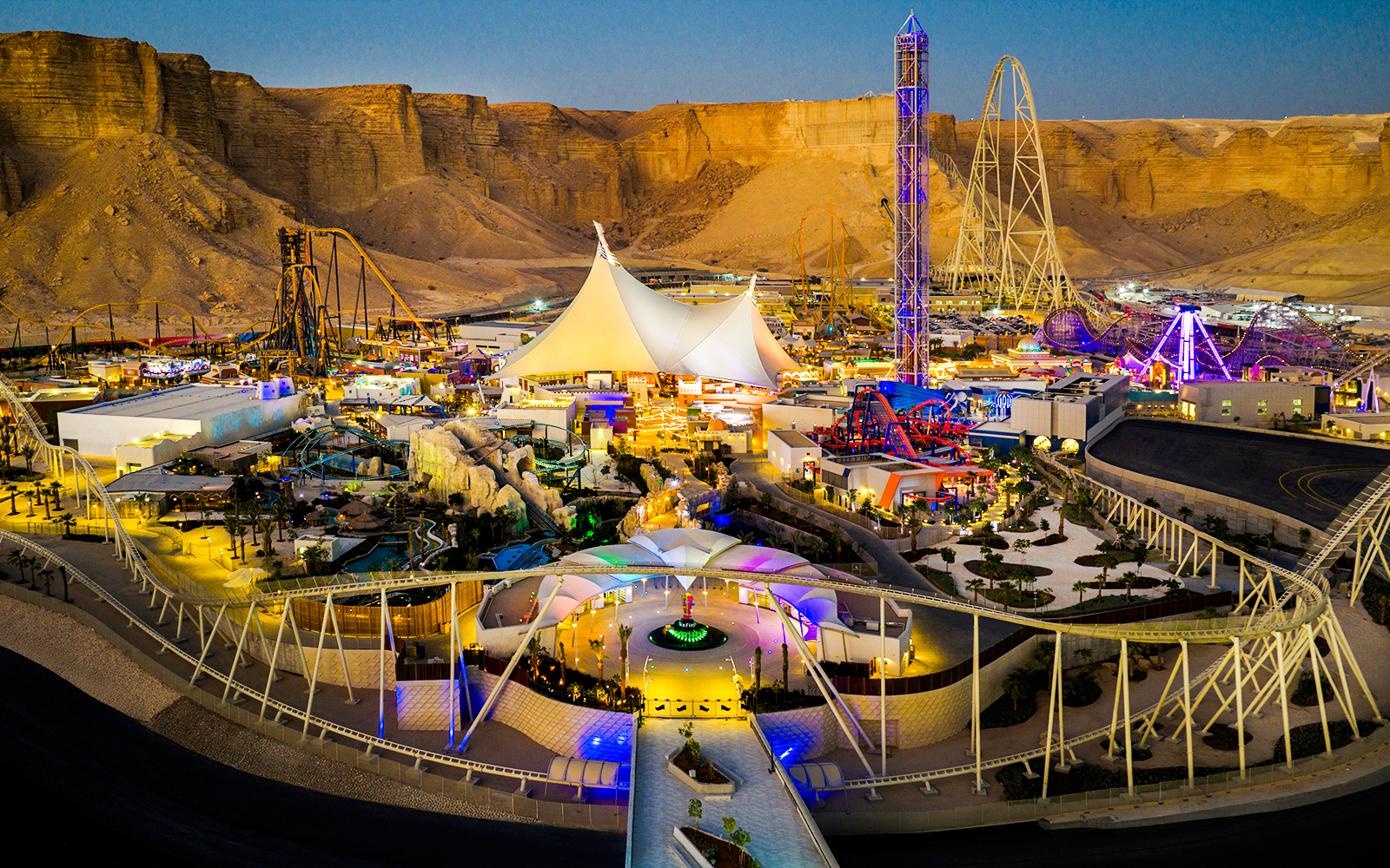 Aerial view of Six Flags Qiddiya City with illuminated rides and attractions against a desert backdrop.