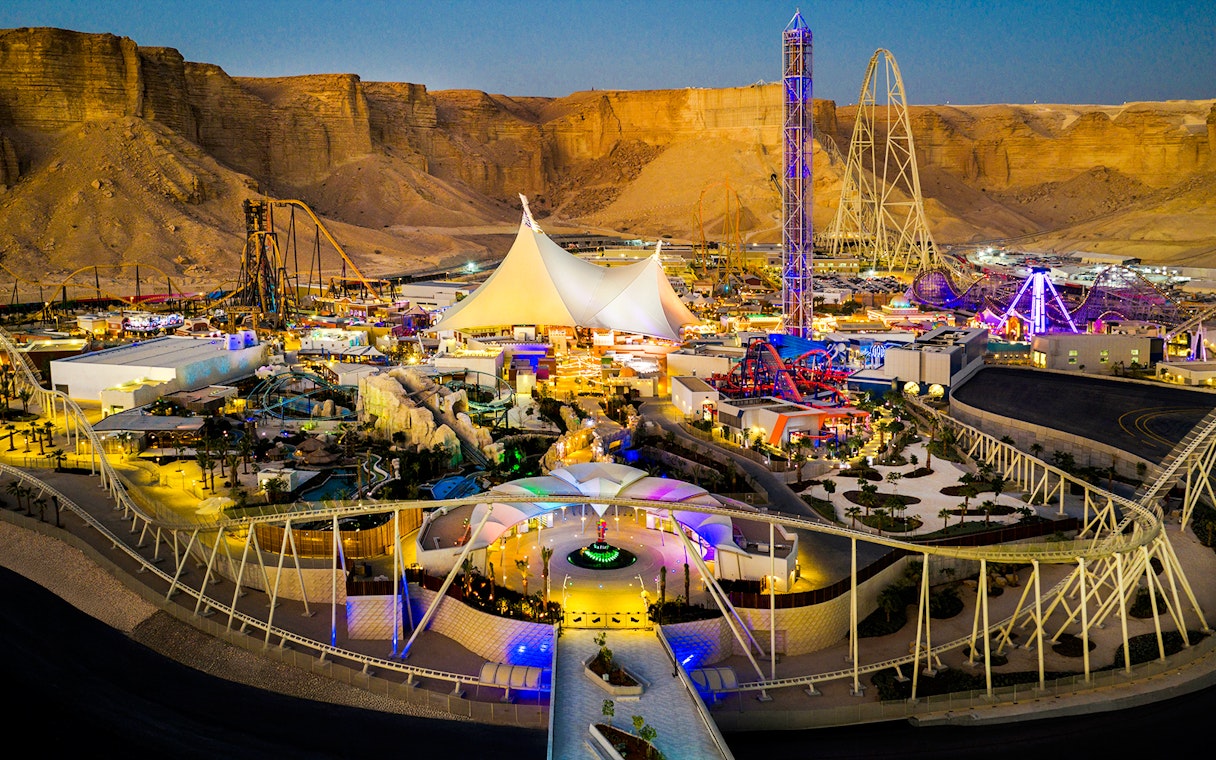 Aerial view of Six Flags Qiddiya City with illuminated rides and attractions against a desert backdrop.