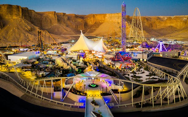 Aerial view of Six Flags Qiddiya City with illuminated rides and attractions against a desert backdrop.