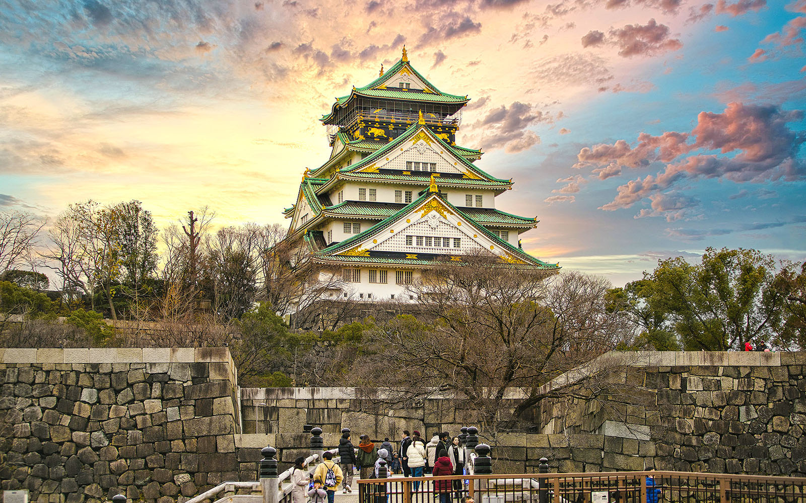 Osaka Castle with sunset sky, surrounded by trees and stone walls, visitors in foreground.
