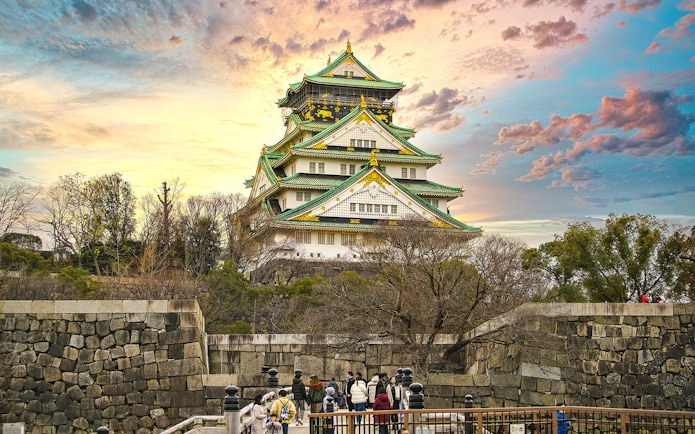 Osaka Castle with sunset sky, surrounded by trees and stone walls, visitors in foreground.