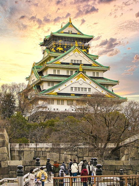 Osaka Castle with sunset sky, surrounded by trees and stone walls, visitors in foreground.