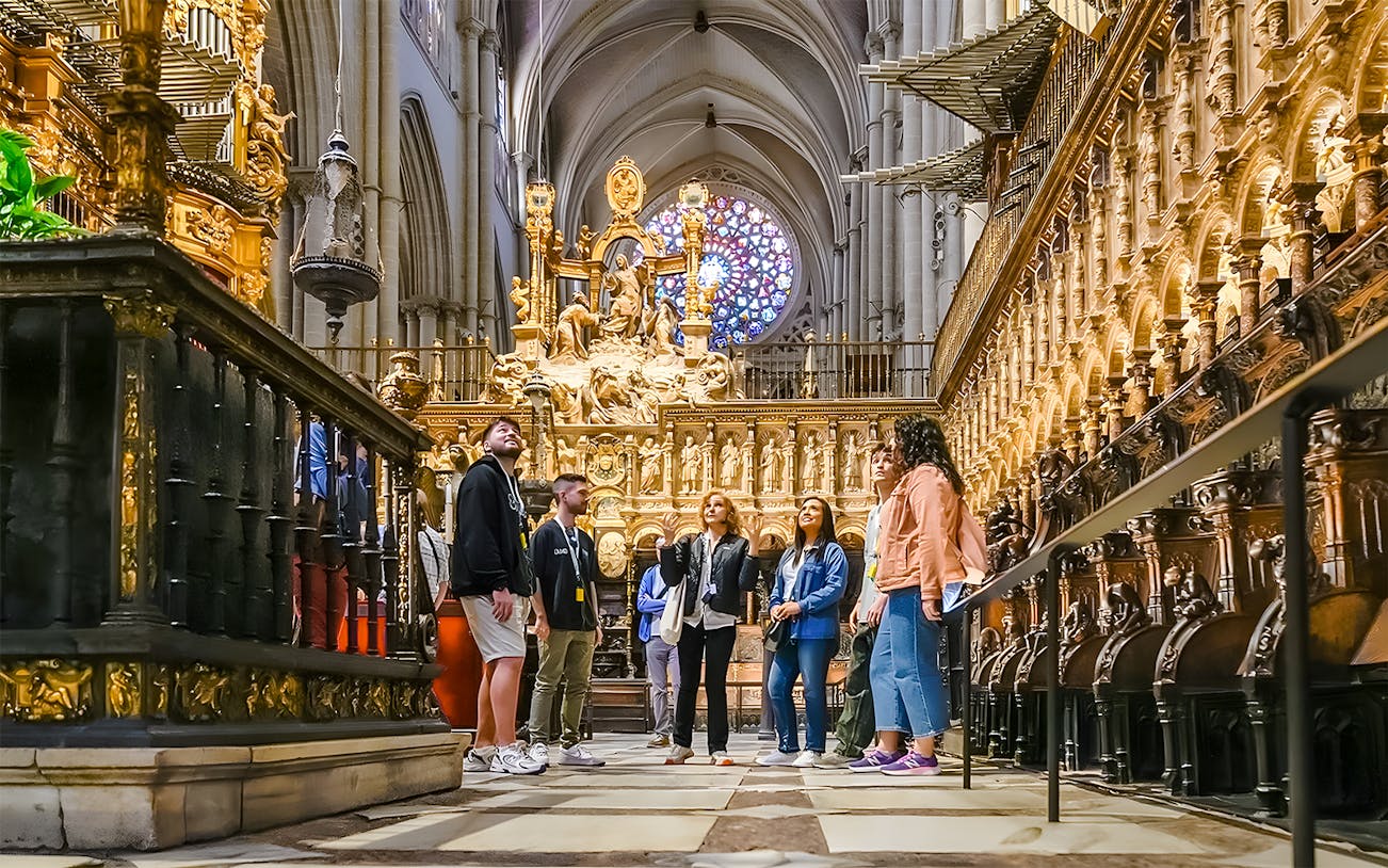 Tour group inside Toledo Cathedral, admiring ornate architecture and stained glass window.