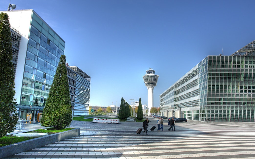 Munich Airport terminal with control tower and travelers walking with luggage.