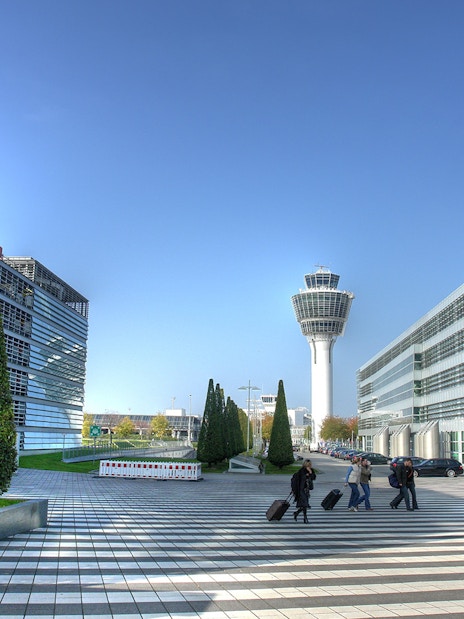 Munich Airport terminal with control tower and travelers walking with luggage.