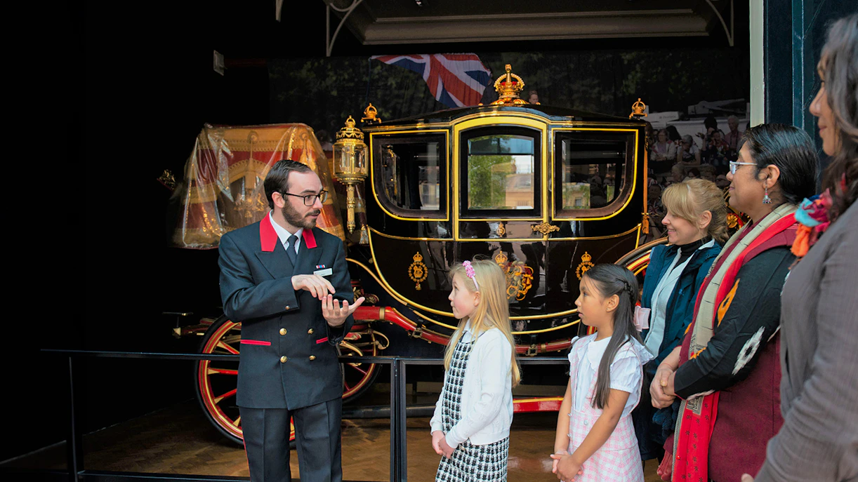 Warden explaining to family visitors in front of a royal carriage at The Royal Mews.