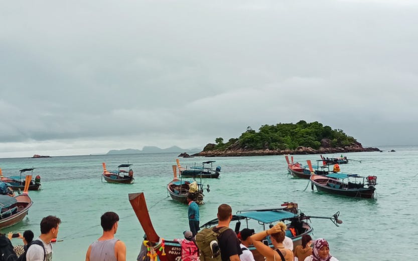 Tourists boarding long-tail boats on a sandy beach in Thailand.
