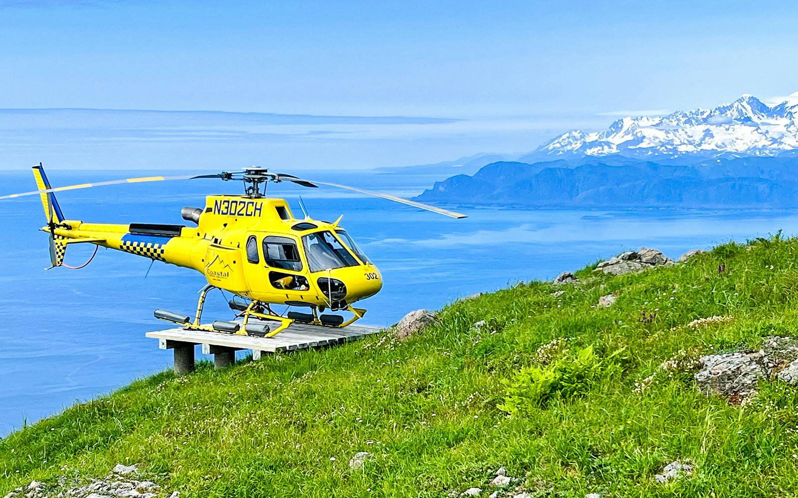 Yellow helicopter on a grassy hill with ocean and mountains in Juneau, Alaska.