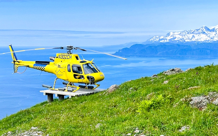 Yellow helicopter on a grassy hill with ocean and mountains in Juneau, Alaska.