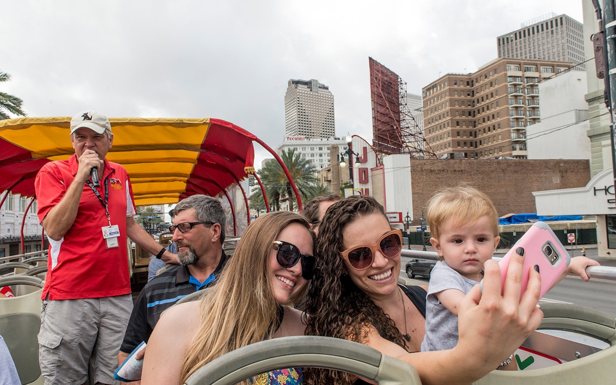 Tourists taking a selfie on a hop-on hop-off bus in New Orleans with city buildings in the background.