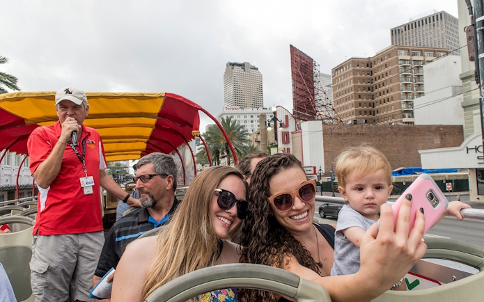 Tourists taking a selfie on a hop-on hop-off bus in New Orleans with city buildings in the background.