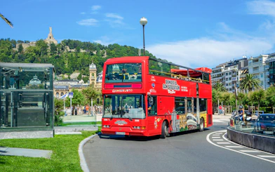 Red double-decker tour bus in San Sebastian with Monte Urgull in the background.