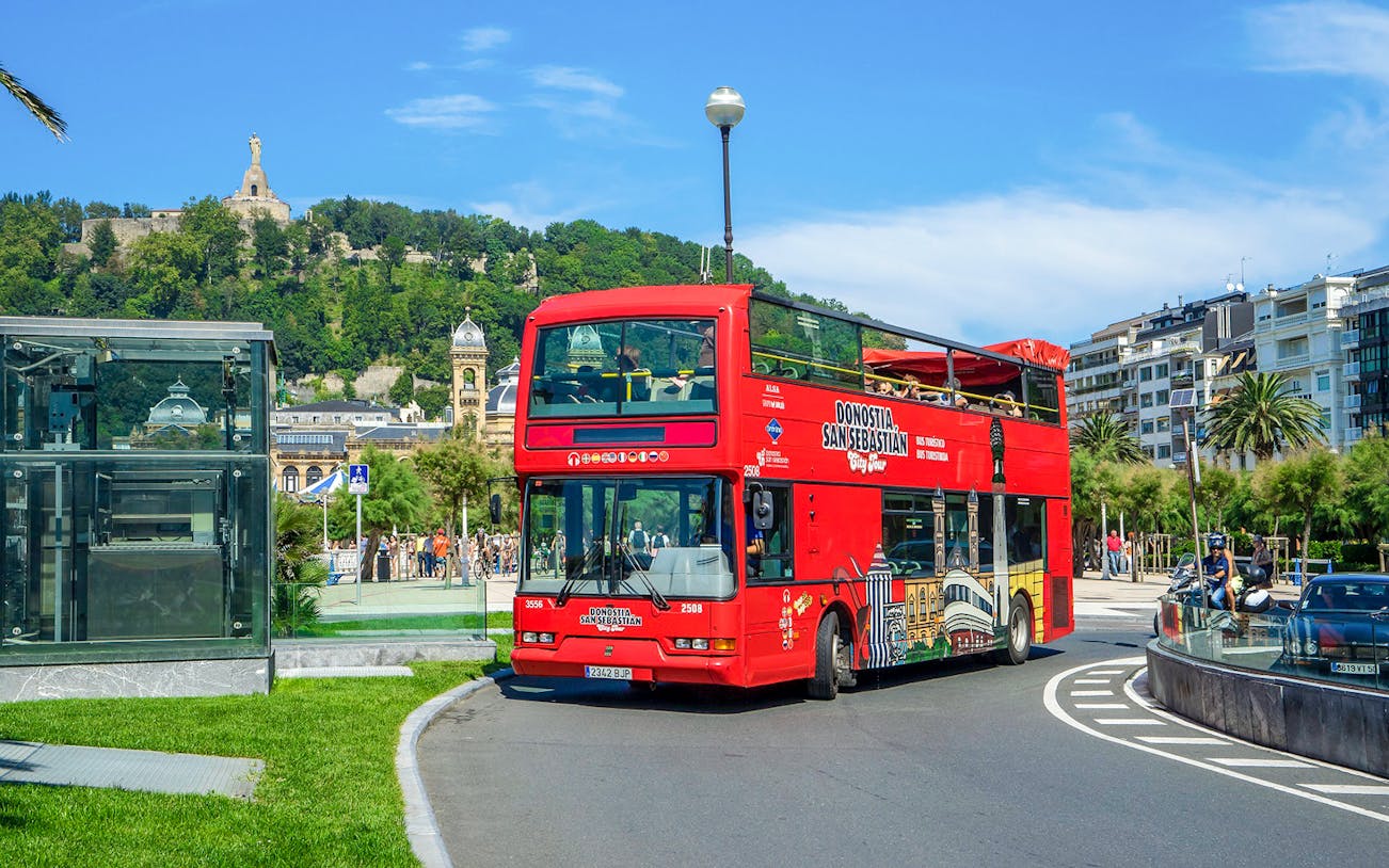 Red double-decker tour bus in San Sebastian with Monte Urgull in the background.