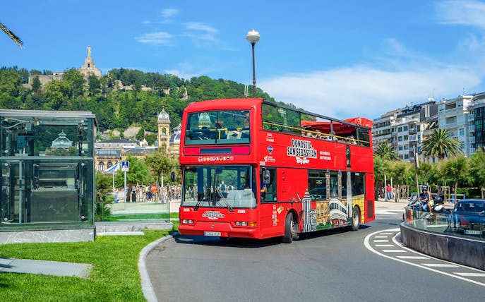 Red double-decker tour bus in San Sebastian with Monte Urgull in the background.