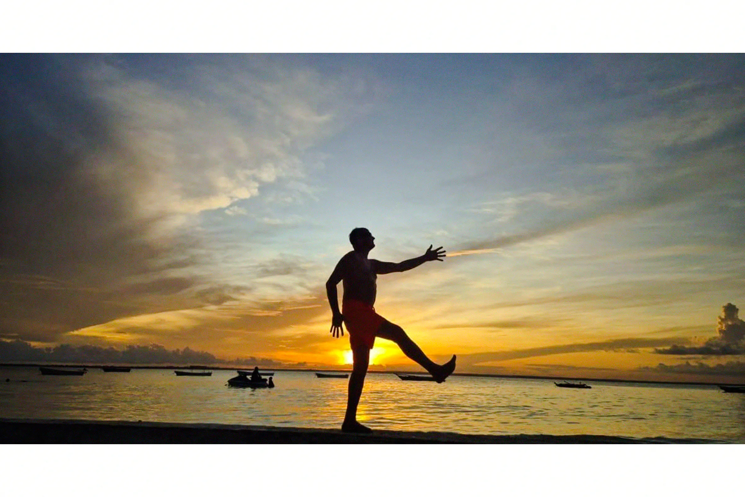 Silhouette of a person on a Zanzibar beach at sunset, with boats in the background.