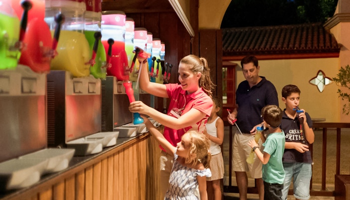 Dining area at Isla Mágica theme park, Seville, Spain, with tables and chairs under a shaded canopy.