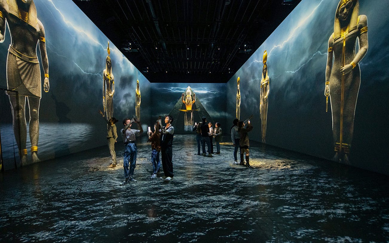 Visitors explore Tutankhamun immersive exhibition with large Egyptian statues projected on walls.