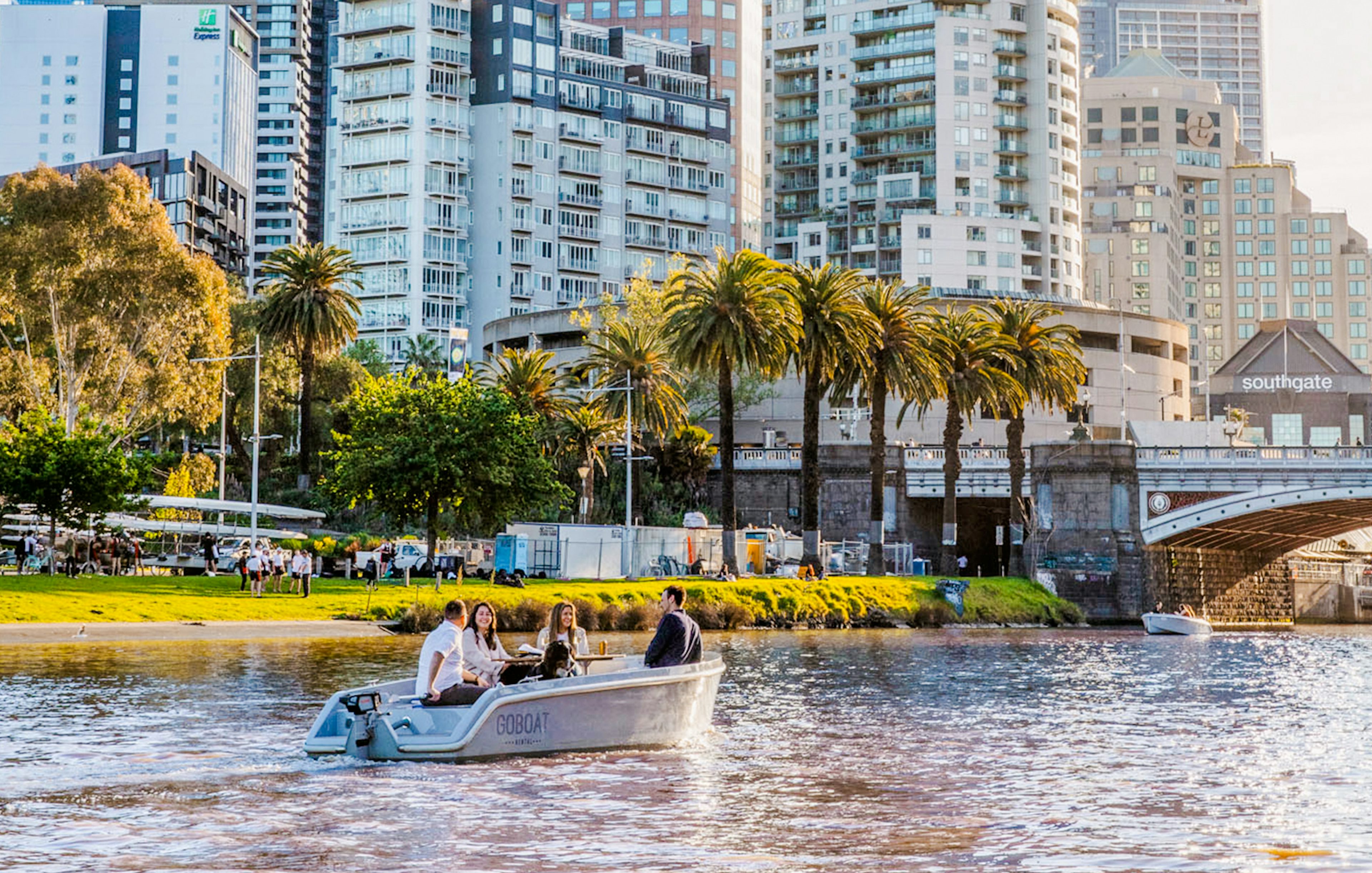 Self-drive boat on Yarra River with cityscape and palm trees in Melbourne.
