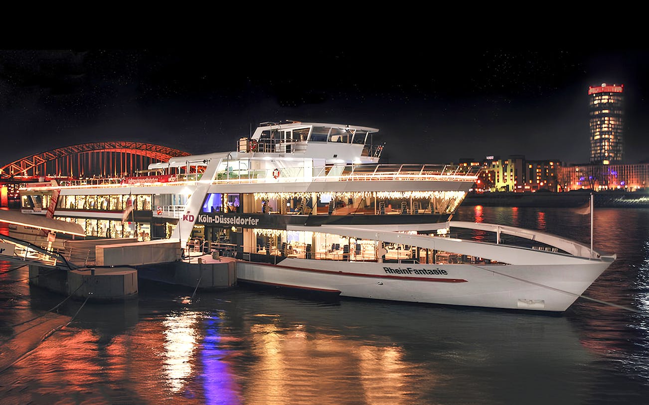 Cruise ship docked at night in Cologne with city lights reflecting on the Rhine River.