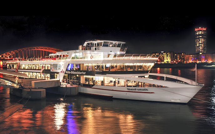 Cruise ship docked at night in Cologne with city lights reflecting on the Rhine River.