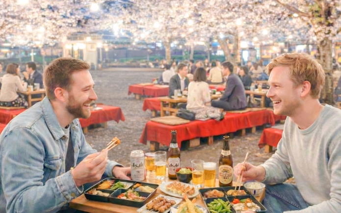 Two friends enjoying drinks under cherry blossoms at night in Kyoto.