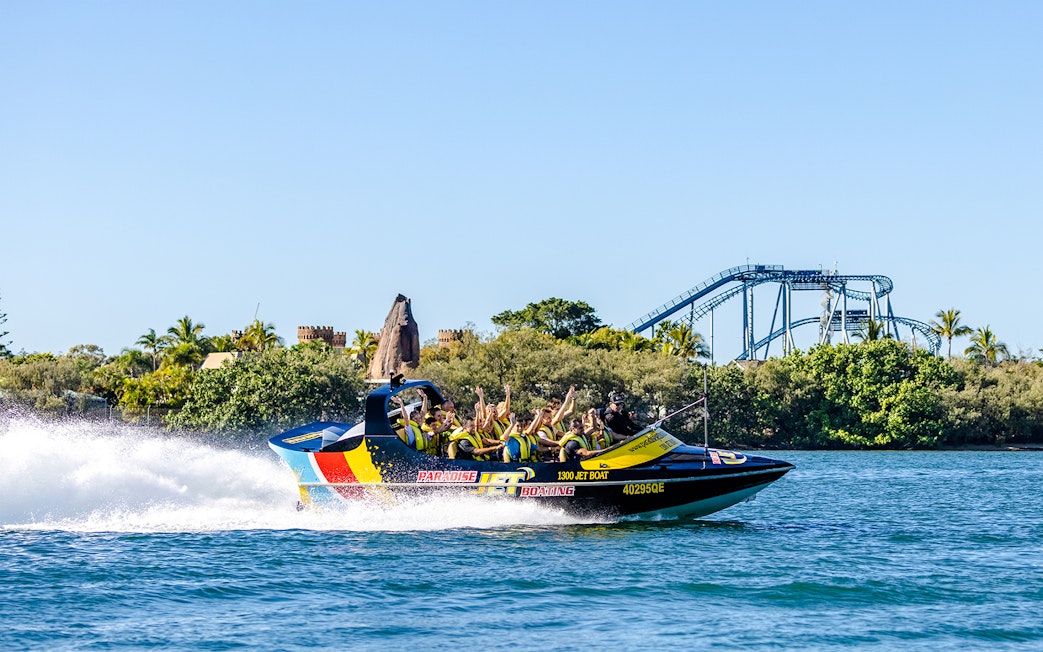 Jet boat with passengers speeding on Gold Coast waterway, roller coaster in background.