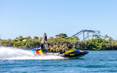 Jet boat with passengers speeding on Gold Coast waterway, roller coaster in background.