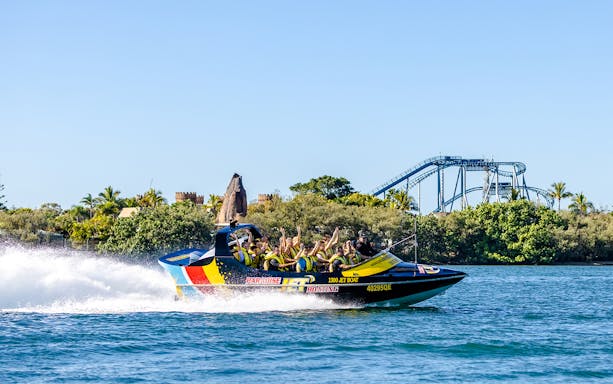 Jet boat with passengers speeding on Gold Coast waterway, roller coaster in background.