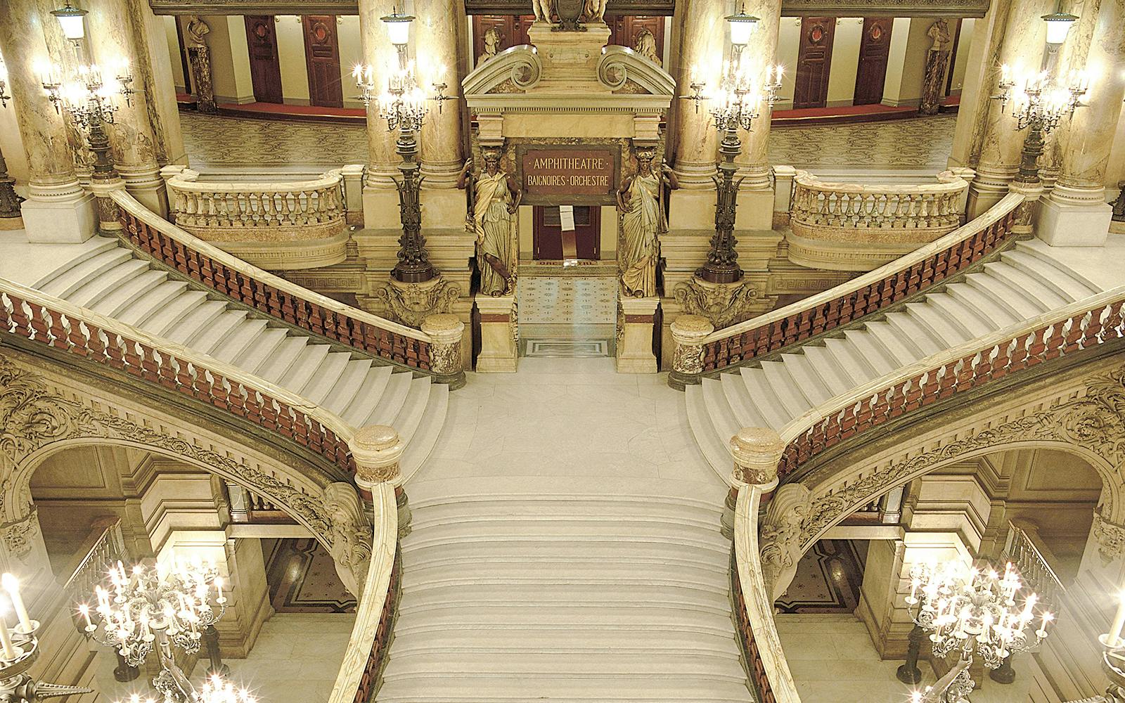 Grand staircase inside Opéra Garnier, Paris, with ornate chandeliers and sculptures.