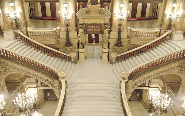 Grand staircase inside Opéra Garnier, Paris, with ornate chandeliers and sculptures.