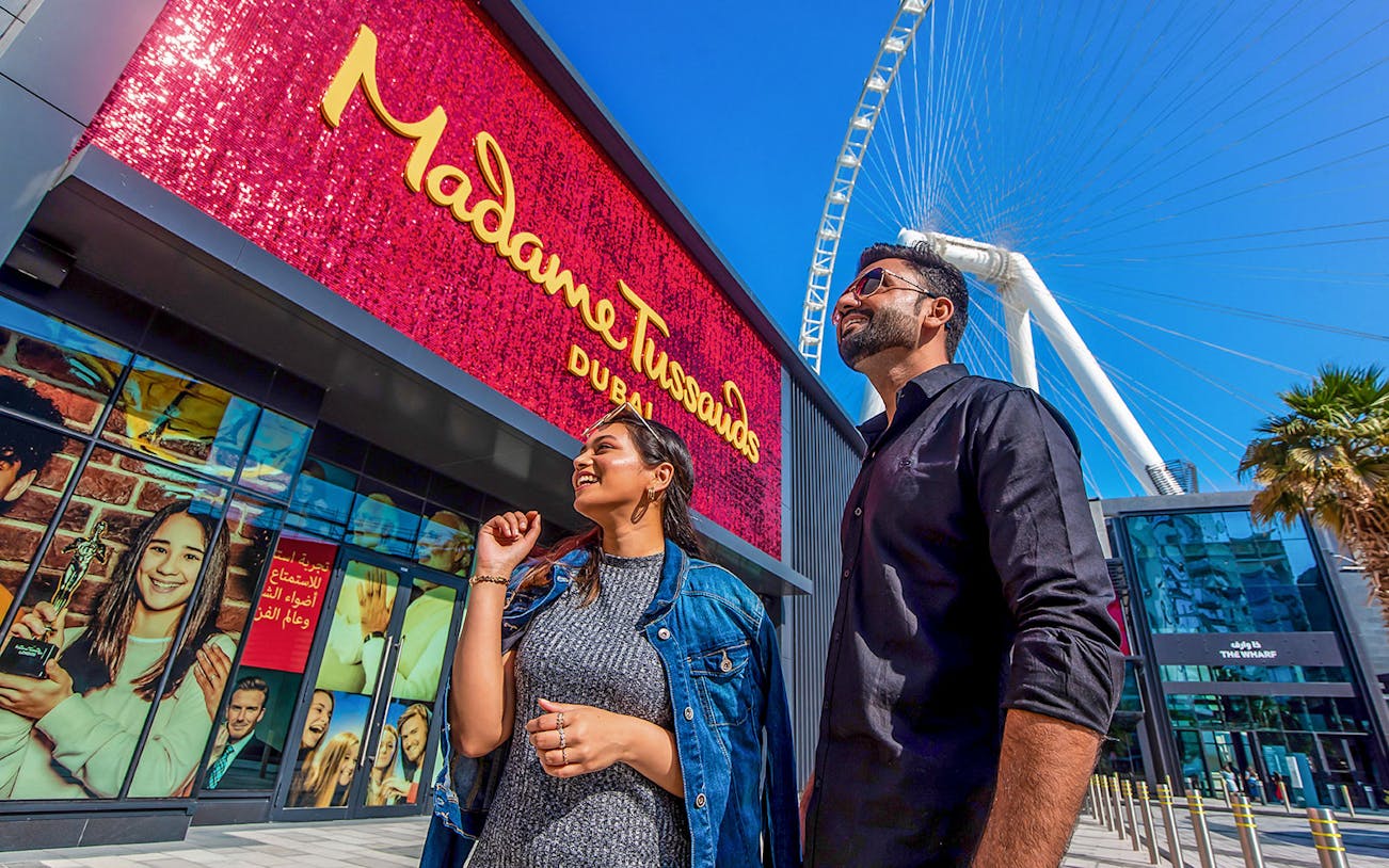 Couple smiling near Madame Tussauds Dubai entrance with Ferris wheel in background.
