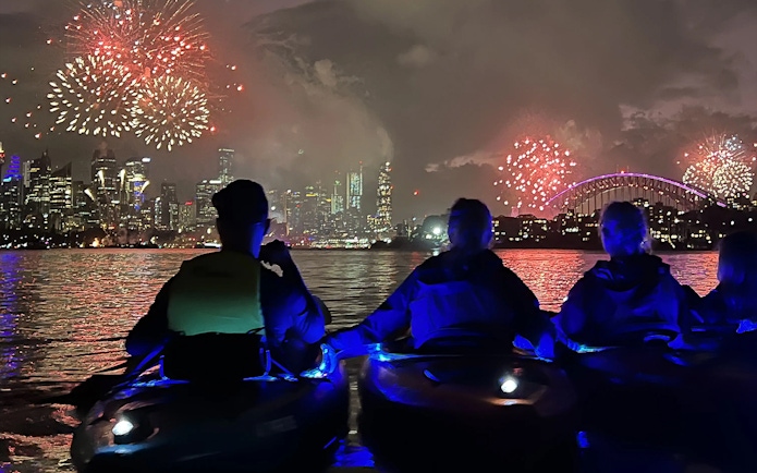 Kayakers in LED-lit boats on Sydney Harbour at dusk with Harbour Bridge and fireworks.