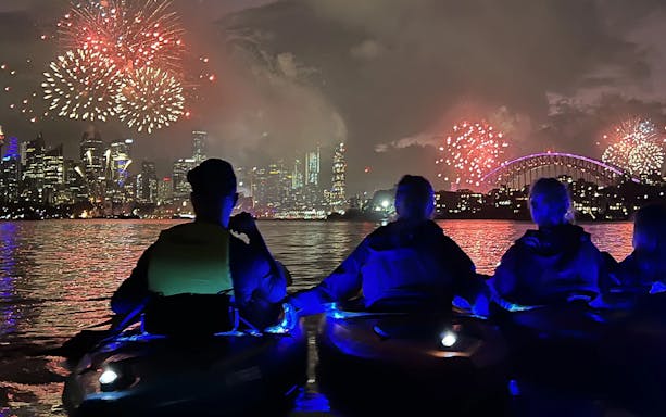Two kayakers in LED-lit boats on Sydney Harbour at dusk with Harbour Bridge illuminated in the background.