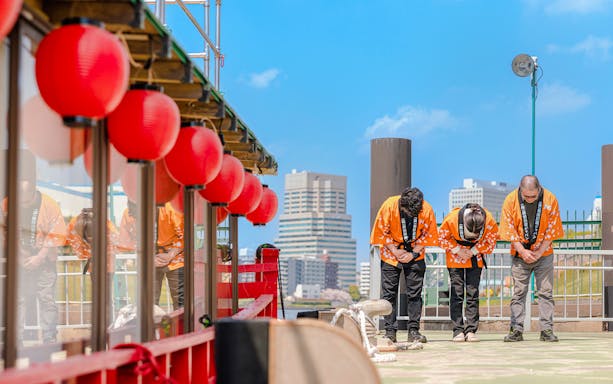 Tour guides bowing on a boat deck with red lanterns in a cityscape background.