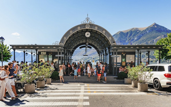 Dock of Bellagio with tourists gathering under an ornate archway, Lake Como in the background.