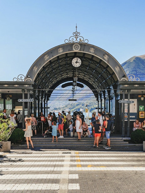 Dock of Bellagio with tourists gathering under an ornate archway, Lake Como in the background.