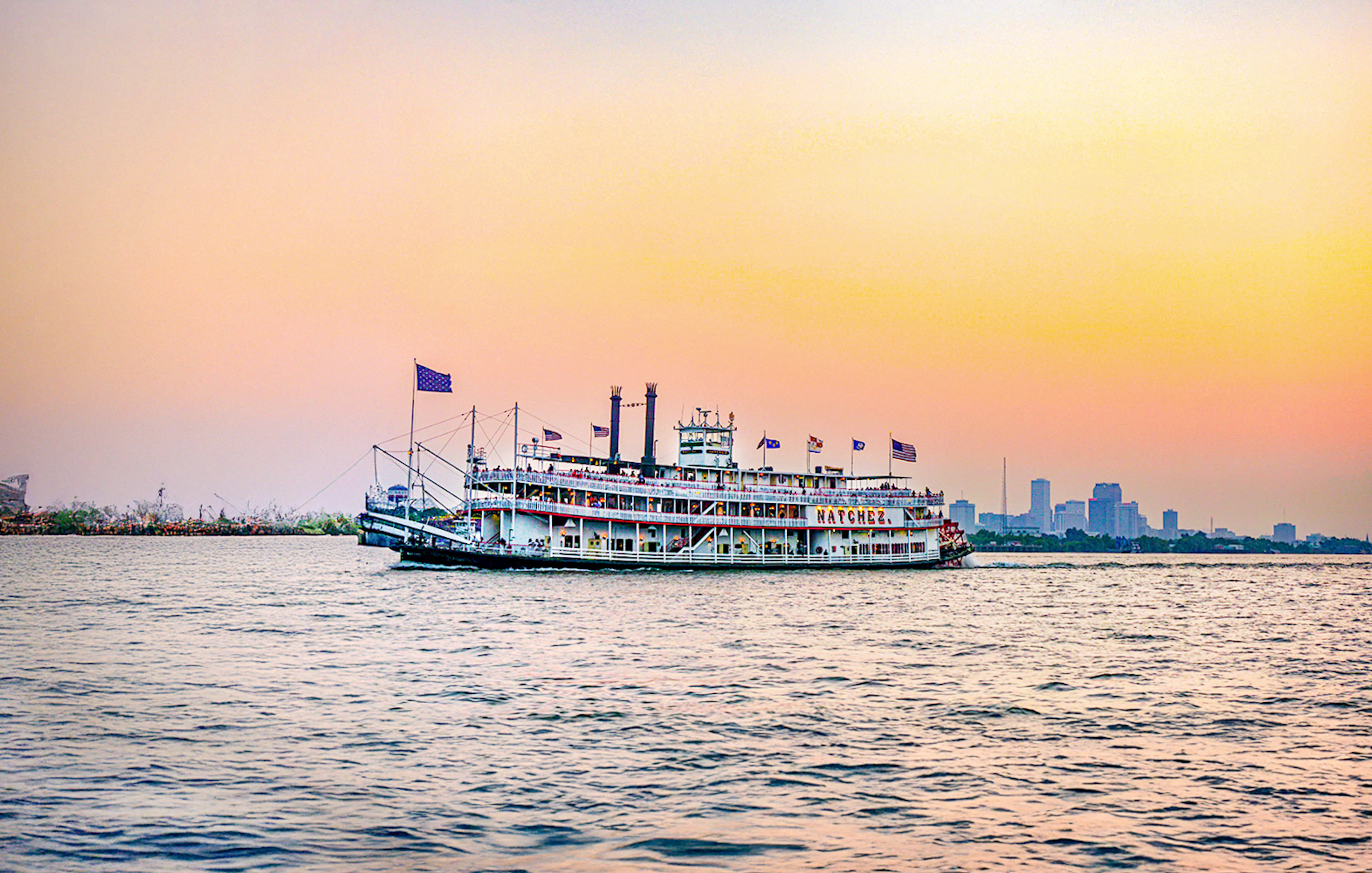 Steamboat Natchez cruising at sunset with city skyline in the background.