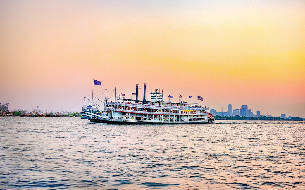 Steamboat Natchez cruising at sunset with city skyline in the background.