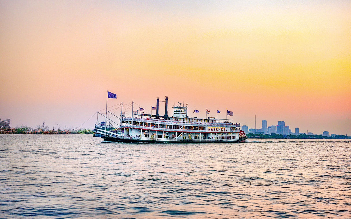 Steamboat Natchez cruising at sunset with city skyline in the background.