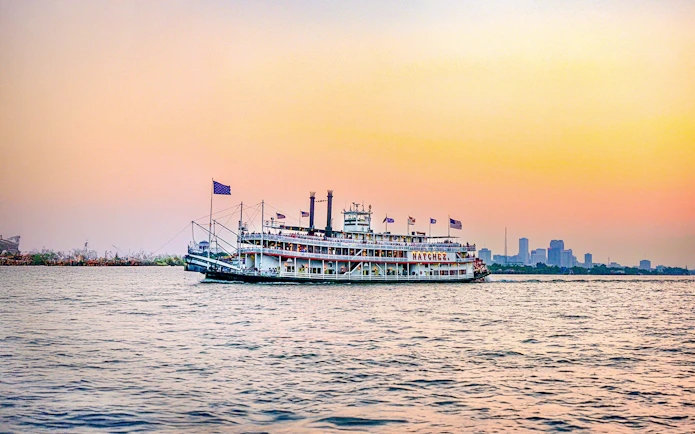 Steamboat Natchez cruising at sunset with city skyline in the background.