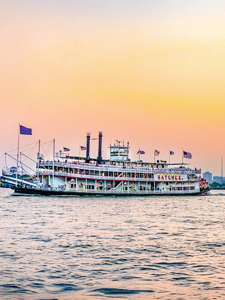 Steamboat Natchez cruising at sunset with city skyline in the background.