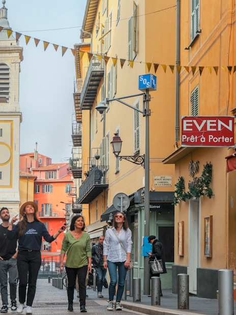 Group walking through colorful street in Nice with bell tower in background.
