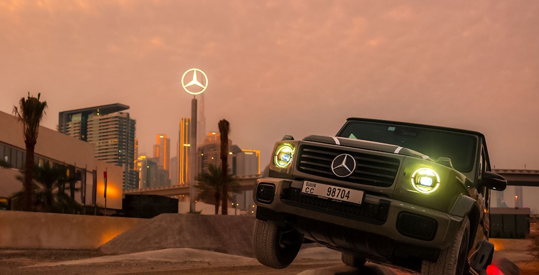 Mercedes-Benz SUV navigating terrain at sunset, Dubai skyline in background.