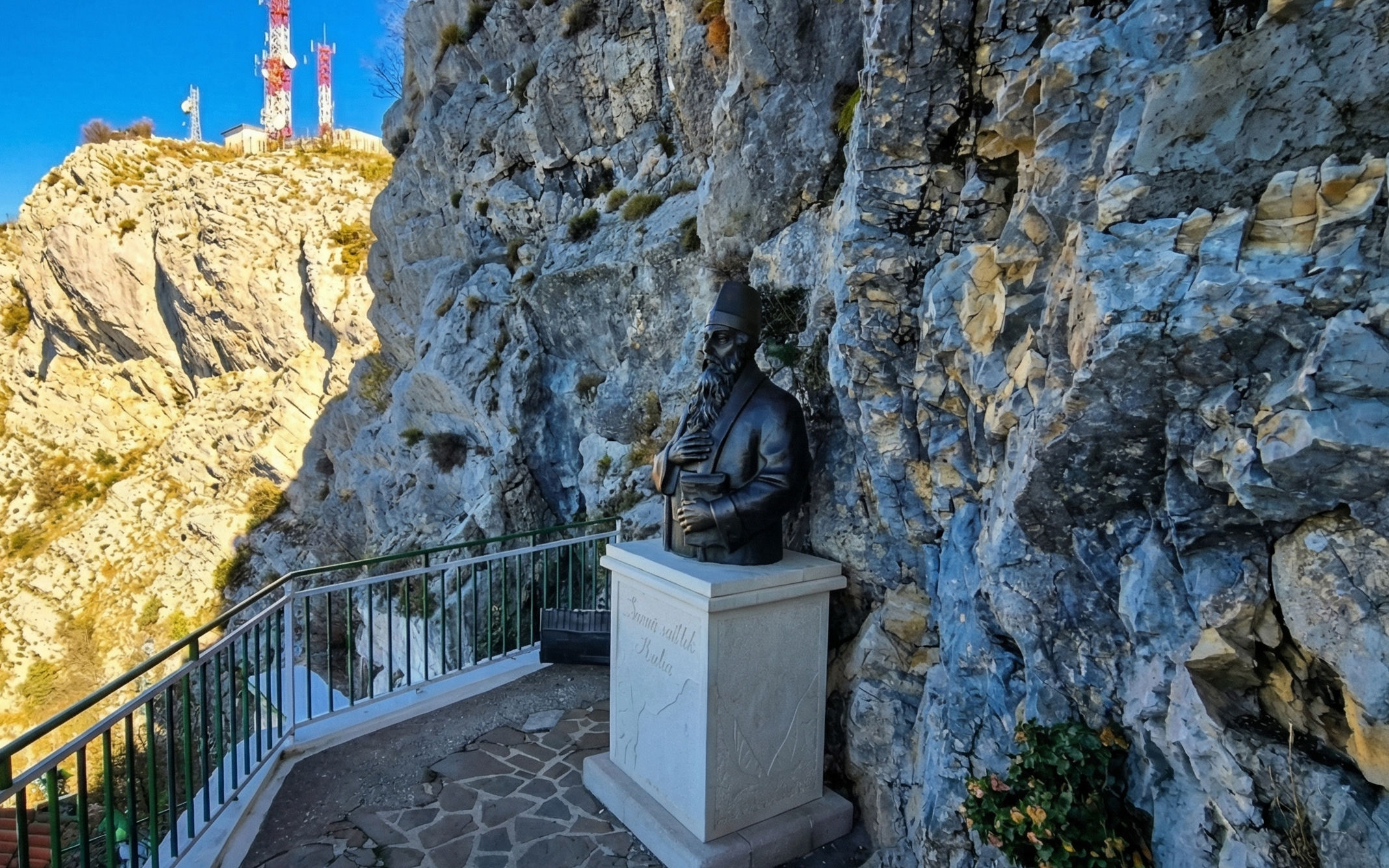 Bust of Sari Saltik in Kruje, Albania, set against a rocky cliff with distant communication towers.