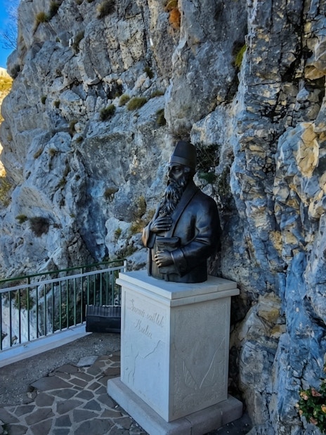 Bust of Sari Saltik in Kruje, Albania, set against a rocky cliff with distant communication towers.