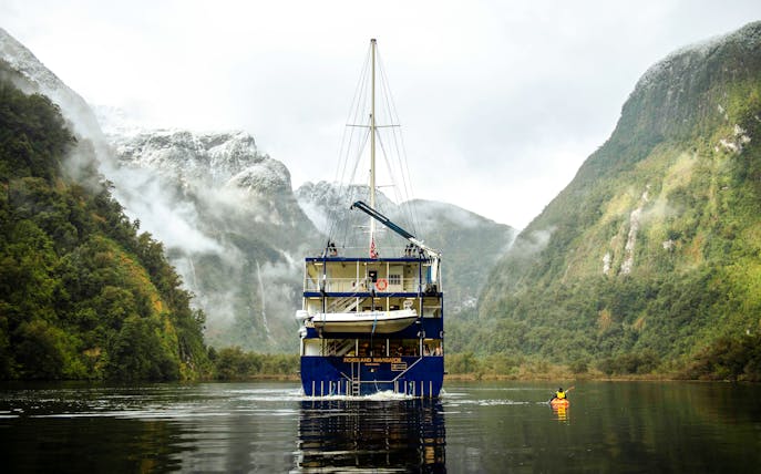 Cruise ship in Doubtful Sound with kayaker and snow-dusted mountains in the background.