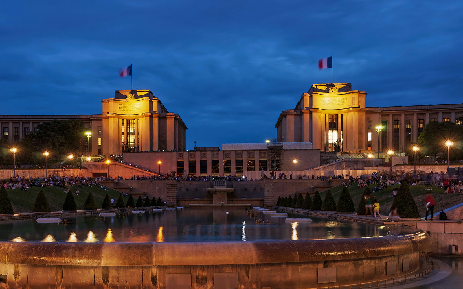 Palais de Chaillot illuminated at night with fountains and people gathered in the foreground, Paris.