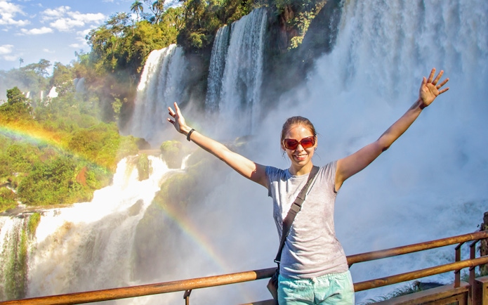 Tourist at Iguazu Falls with arms raised, rainbow visible.