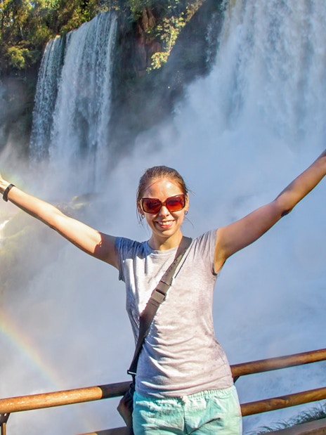 Tourist at Iguazu Falls with arms raised, rainbow visible.