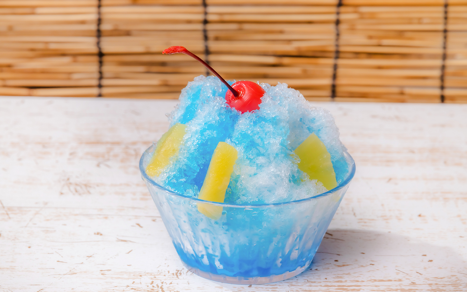 Shaved ice with blue syrup, pineapple slices, and a cherry on top in a glass bowl.